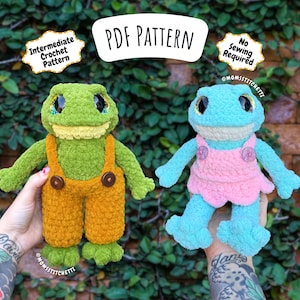 Peut inclure: Deux peluches en forme de grenouille crochetées, une verte avec une salopette orange et l'autre bleue avec une robe rose. L'image comprend le texte "PDF Pattern", "Intermediate Crochet Pattern" et "No Sewing Required".