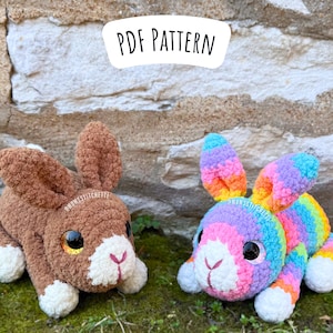 以下が含まれることがあります： 2つの編みぐるみウサギ、1つは茶色、もう1つは虹色のストライプ。茶色のウサギは顔と足に白いアクセントがあります。虹色のウサギは顔と足に白いアクセントがあります。ウサギは石の壁の前にある緑の苔のベッドの上に座っています。ウサギの上に「PDF PATTERN」というテキストが表示されています。