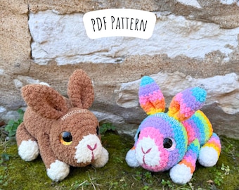 Op de afbeelding: Twee gehaakte konijnenknuffels, een bruin en een met regenboogstrepen. Het bruine konijn heeft witte accenten op zijn gezicht en poten. Het regenboogkonijn heeft witte accenten op zijn gezicht en poten. De konijnen zitten op een bed van groen mos voor een stenen muur. De tekst "PDF PATTERN" wordt boven de konijnen weergegeven.