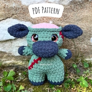 May include: A crocheted plush toy in the shape of a monster with green and blue fur, pink brain, and red stitches. The toy has large black eyes and a stitched-on mouth. The toy is sitting on a bed of green grass and a stone wall. The text "PDF PATTERN" is written in white on a black circle above the toy.