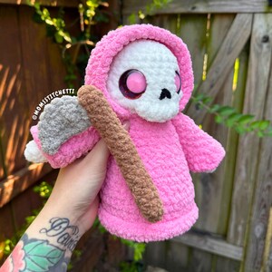 May include: A crocheted plush figure featuring a white skull head with large pink eyes, wearing a bright pink hooded robe. The figure holds a gray and brown scythe. The text @MOMSSTITCHETTI is visible on the side of the plush figure's hood.