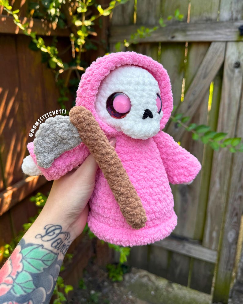 May include: A crocheted plush figure featuring a white skull head with large pink eyes, wearing a bright pink hooded robe. The figure holds a gray and brown scythe. The text @MOMSSTITCHETTI is visible on the side of the plush figure's hood.