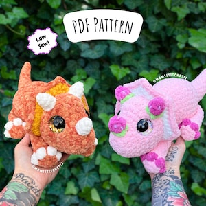 May include: Two crocheted triceratops dinosaurs, one orange and one pink, are displayed. The orange dinosaur has white horns and accents, while the pink one has green and purple accents. The image includes text that reads "PDF PATTERN" and "Low Sew!"
