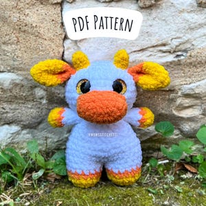 Puede incluir: Una vaca de peluche blanca tejida a crochet con detalles en naranja y amarillo. La vaca tiene ojos grandes y brillantes y una nariz naranja esponjosa. El texto "PDF PATTERN" es visible sobre la vaca.