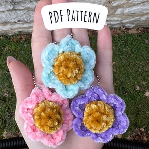 May include: Three crocheted flower keychains in pink, blue, and purple with a gold center. The keychains are hanging from a silver chain. The text "PDF PATTERN" is written in white on a black background.