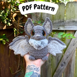 May include: A gray crocheted bat plush toy with large, dark, reflective eyes, pointed ears with lighter gray inner sections, and wings featuring light gray edges. The chest has a patch of fluffy, textured gray fur. Text reads PDF Pattern.