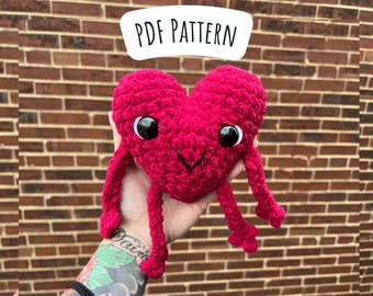 May include: A crocheted heart-shaped plush toy with long arms and legs. The toy is pink and has black eyes and a smiling mouth.