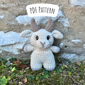 May include: A crocheted goat amigurumi toy with brown horns, light brown fur, and large black eyes. The toy is sitting on a bed of green moss and brown leaves. The text "PDF PATTERN" is written in white on a white background.