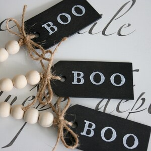 May include: Three black chalkboard tags with the word "BOO" written in white paint. The tags are tied together with twine and strung with a strand of white wooden beads.