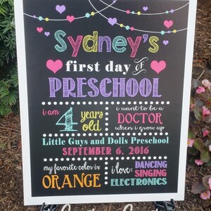 Puede incluir: Un cartel de pizarra colorido con el texto "Sydney's first day of PRESCHOOL" y detalles sobre la edad del ni&ntilde;o, su color favorito y sus intereses. El cartel est&aacute; decorado con corazones, estrellas y una cadena de luces.