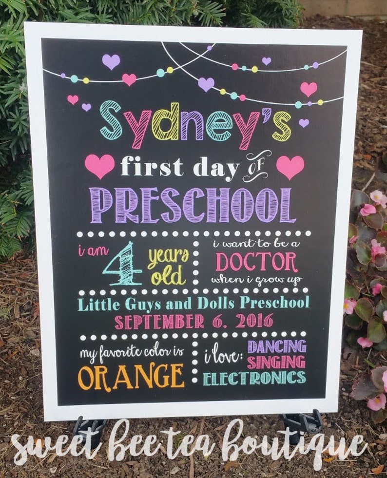 Puede incluir: Un cartel de pizarra colorido con el texto "Sydney's first day of PRESCHOOL" y detalles sobre la edad del ni&ntilde;o, su color favorito y sus intereses. El cartel est&aacute; decorado con corazones, estrellas y una cadena de luces.