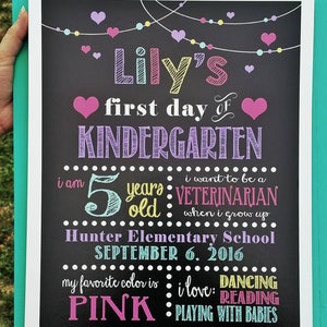 Puede incluir: Un letrero de pizarra con el texto "Lily's first day of KINDERGARTEN" en morado y rosa. El letrero tambi&eacute;n incluye detalles sobre la edad de Lily, su color favorito y sus aspiraciones profesionales futuras. El letrero est&aacute; decorado con corazones coloridos y luces de cadena.