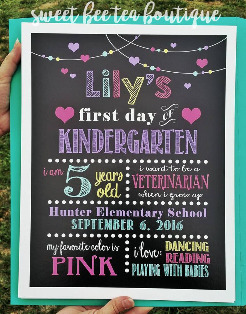 Puede incluir: Un letrero de pizarra con el texto "Lily's first day of KINDERGARTEN" en morado y rosa. El letrero tambi&eacute;n incluye detalles sobre la edad de Lily, su color favorito y sus aspiraciones profesionales futuras. El letrero est&aacute; decorado con corazones coloridos y luces de cadena.