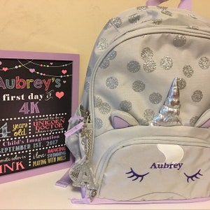 Puede incluir: Una mochila gris de unicornio con lunares plateados de purpurina y un cuerno de unicornio morado. La mochila tiene un bolsillo con cremallera y un bolsillo de malla en el lateral. La mochila tiene el nombre "Aubrey" bordado en la parte delantera. Un letrero de pizarra con el texto "Aubrey's first day of 4K" est&aacute; en el fondo.