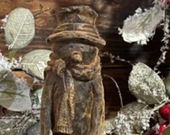 May include: A wooden snowman figure, featuring a top hat, scarf, and a wreath. The snowman is painted in brown tones with a distressed finish. The background includes decorative leaves and berries. The figure has a vintage appearance.