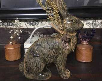 May include: A ceramic rabbit figurine with a distressed, mottled brown and green finish. The rabbit has dried floral accents and a small bell around its neck. The figurine is displayed on a dark wooden surface.