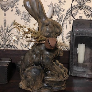May include: A dark brown rabbit figurine with a bell around its neck and a bundle of dried flowers. The rabbit is sitting on a base and has a feather in its ear. A decorative lantern and a small box are in the background.