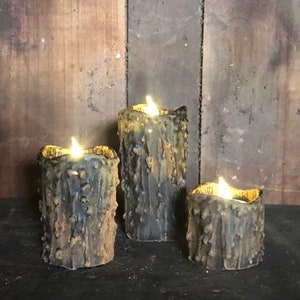 May include: Three battery-operated, rustic-style candles with flickering LED lights. The candles have a textured, weathered appearance in shades of brown, blue, and gold. The candles are of varying heights, creating a layered effect, and are set against a dark background.