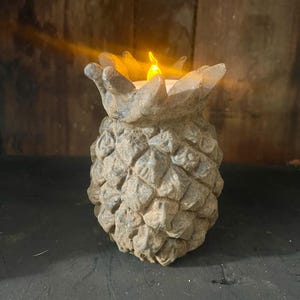May include: A decorative, weathered-looking pineapple-shaped candle holder with a flickering yellow light. The candle holder is made of a textured, gray-brown material and sits on a dark surface. The light resembles a candle flame.