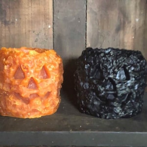 May include: Two Halloween-themed candle holders. One is orange and the other is black. Both are shaped like jack-o'-lanterns with carved faces. The candle holders are sitting on a dark surface against a wooden background.