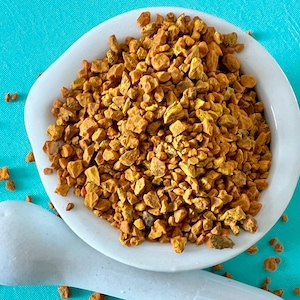 May include: A white ceramic mortar filled with small, golden-brown pieces of a spice, possibly turmeric or a similar herb. The mortar is on a bright turquoise surface, with a white pestle in the foreground.