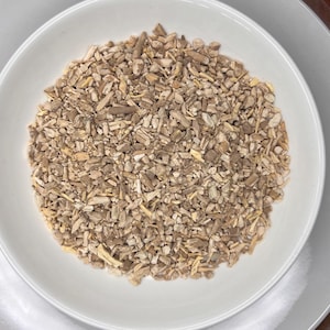 May include: A white ceramic bowl filled with dried, chopped herbs and spices. The bowl is on a white plate, which is on a wooden tray. The herbs and spices are various shades of brown and beige.