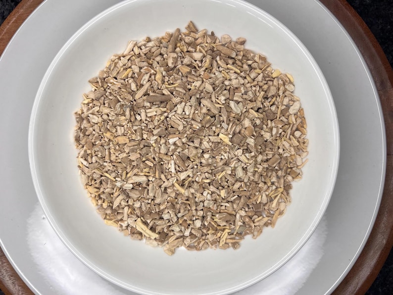 May include: A white ceramic bowl filled with dried, chopped herbs and spices. The bowl is on a white plate, which is on a wooden tray. The herbs and spices are various shades of brown and beige.
