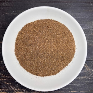 May include: A white ceramic plate filled with a mound of brown, finely ground spice. The spice has a uniform texture and color, and the plate is set against a dark wooden surface.
