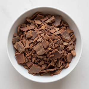 May include: A white ceramic bowl filled with pieces of brown cinnamon bark. The bark pieces vary in size and shape, with a rough, textured appearance. The bowl is set against a white background.