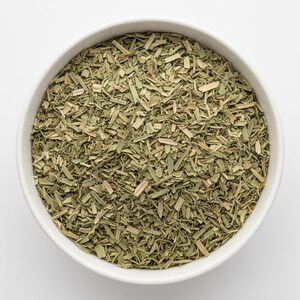 May include: A white ceramic bowl filled with dried, finely chopped green lemongrass stalks. The herb pieces are a mix of pale tan and muted sage green, showing a fibrous, shredded texture throughout the bowl, viewed from directly above.