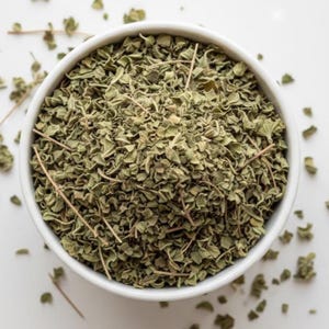 May include: Overhead shot of a white bowl filled with dried oregano leaves. The leaves are a muted green color, with some stems visible. The bowl is set against a white background, with a few leaves scattered around.