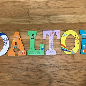 May include: Wooden letters spelling "DALTON" are arranged on a wood floor. Each letter is a different color and decorated with science-themed illustrations, including molecules, beakers, and a microscope. The letters are painted in bright colors.