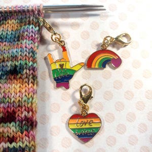 May include: A close-up of three colorful enamel charms with gold-colored clasps. One charm is a rainbow-colored hand making the 'I love you' sign. Another is a rainbow with a heart. The third is a rainbow heart with the words "Love Wins".