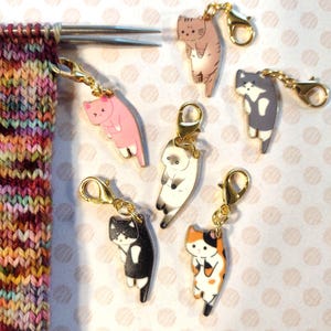 May include: A collection of cat-shaped stitch markers in various colors, including pink, brown, black, and white, each with a gold-colored clasp. The markers are attached to knitting needles and a colorful knitted fabric.