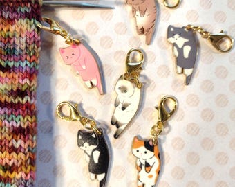 Puede incluir: Una colección de marcadores de punto con forma de gato en varios colores, incluyendo rosa, marrón, negro y blanco, cada uno con un cierre dorado. Los marcadores están unidos a agujas de tejer y a una tela de punto colorida.