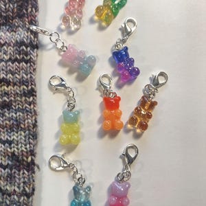 Könnte beinhalten: Eine Sammlung bunter Gummibärchen-Anhänger mit silberfarbenen Karabinerverschlüssen. Die Anhänger sind in verschiedenen Farben erhältlich, darunter Rosa, Blau, Grün, Orange und Gelb. Die Anhänger sind an einem Strickstoff befestigt.