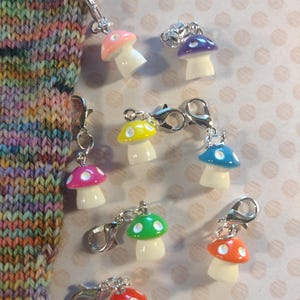 Puede incluir: Una colección de marcadores de punto con forma de seta de colores, con cierres plateados. Las setas tienen sombreros rojos, rosas, naranjas, verdes, amarillos, azules y morados. Los marcadores están unidos a agujas de tejer y a una tela tejida.