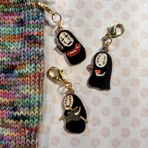 May include: Three black enamel stitch markers with gold-colored clasps, each featuring a character design with a white face and red mouth. A knitted sock in various colors is in the background, with knitting needles. The markers are for knitting and crochet.