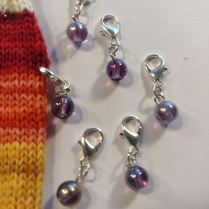 Puede incluir: Una colección de seis colgantes con cierre de mosquetón plateados, cada uno con una cuenta púrpura translúcida. Los colgantes están dispersos sobre una superficie clara, con una porción de un artículo de punto colorido visible en el lado izquierdo.