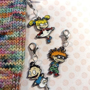 May include: Three cartoon character stitch markers with lobster claw clasps. The characters are Angelica, Tommy, and Chuckie from the animated series. The markers are attached to knitting needles with colorful yarn.