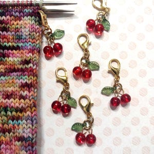 May include: Gold-colored stitch markers with red cherry beads and green leaf accents. The stitch markers are attached to a colorful knitted project with metal needles. The cherries are made of translucent red beads.