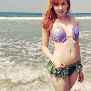Könnte beinhalten: Eine Frau mit roten Haaren trägt ein lilafarbenes Muschel-Bikini-Oberteil und ein grünes, gekräuseltes Bikini-Unterteil am Strand.