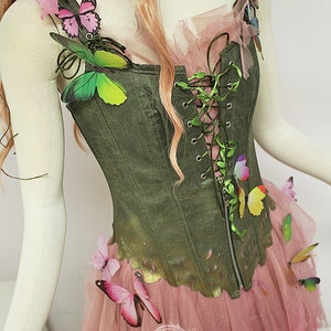 May include: A green corset top with a zip and lace-up detail, decorated with colourful butterfly accents. The top is paired with a pink tulle skirt, creating a whimsical fairy-like aesthetic. The outfit is displayed on a mannequin.