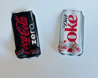 May include: Two soda can-shaped objects. One is black with red "Coca-Cola Zero" text. The other is white with red "Diet Coke" text. Both have small, round "Coke" buttons attached.