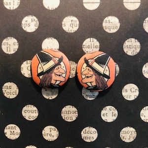 May include: Pair of round button earrings featuring a cartoon illustration of a character with a hat and a smiling face. The earrings have an orange background and are set against a black background with white polka dots.