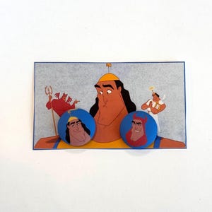 May include: Two blue button pins with images of a cartoon character from the Disney movie Hercules. The character is shown with a red devil horn on his head and a red devil costume. The background of the pins is blue. The pins are on a white background.