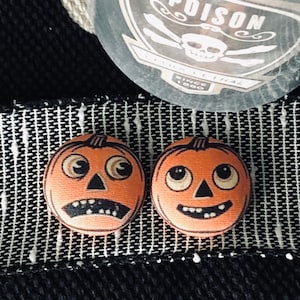 May include: Two orange fabric pumpkin buttons with black eyes, a black nose, and a black mouth. One pumpkin has a wide smile, and the other has a slightly open mouth with teeth.