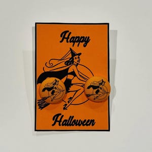 May include: Orange Halloween-themed card with the words "Happy Halloween" in black script. Two round earrings with a witch on a broomstick are attached to the card. The earrings have an orange background and a black witch design.