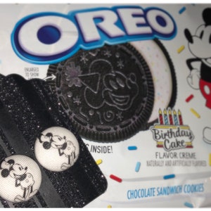 Pode incluir: Um par de brincos de bot&atilde;o Mickey Mouse preto e branco em um fundo preto com glitter. Os brincos est&atilde;o em uma etiqueta com o texto "ENLARGED TO SHOW" e um biscoito Oreo preto e branco com um design de Mickey Mouse. O fundo &eacute; uma embalagem de biscoito Oreo branca com um design de confete vermelho e amarelo e um desenho animado do Mickey Mouse.
