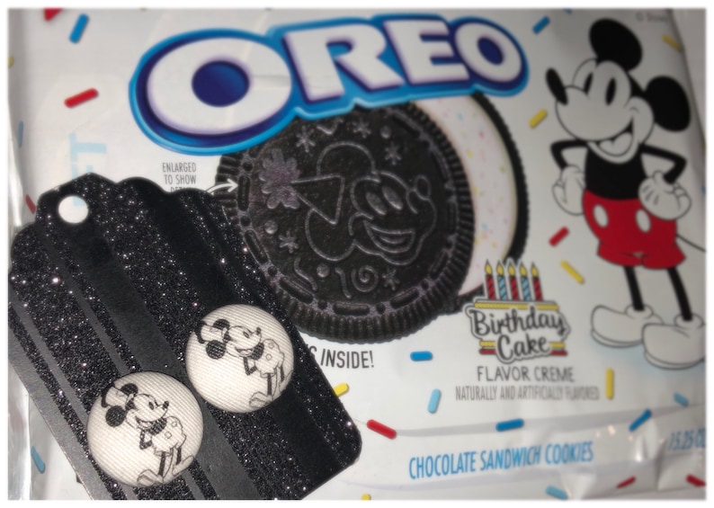 Pode incluir: Um par de brincos de bot&atilde;o Mickey Mouse preto e branco em um fundo preto com glitter. Os brincos est&atilde;o em uma etiqueta com o texto "ENLARGED TO SHOW" e um biscoito Oreo preto e branco com um design de Mickey Mouse. O fundo &eacute; uma embalagem de biscoito Oreo branca com um design de confete vermelho e amarelo e um desenho animado do Mickey Mouse.
